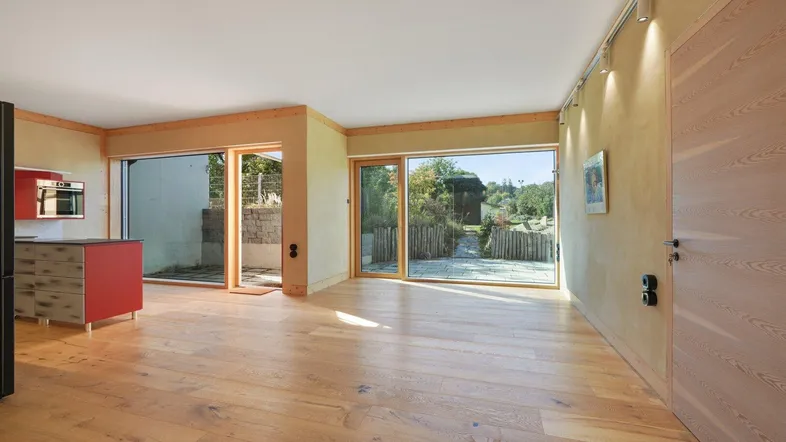 Heller offener Wohnbereich mit Holzboden, integrierter Küche und großen Fenstern zum Garten.