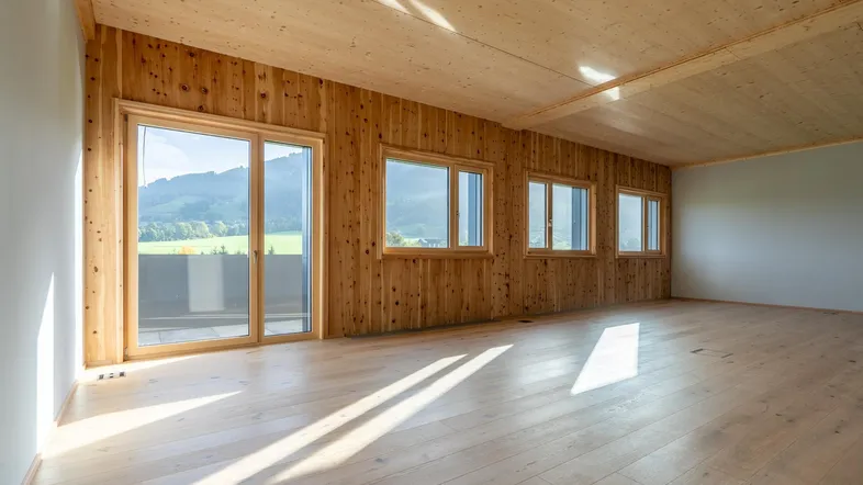 Heller, unmöblierter Raum mit Holzwänden und -decke, großen Fenstern und Zugang zu einem Balkon mit Bergblick.