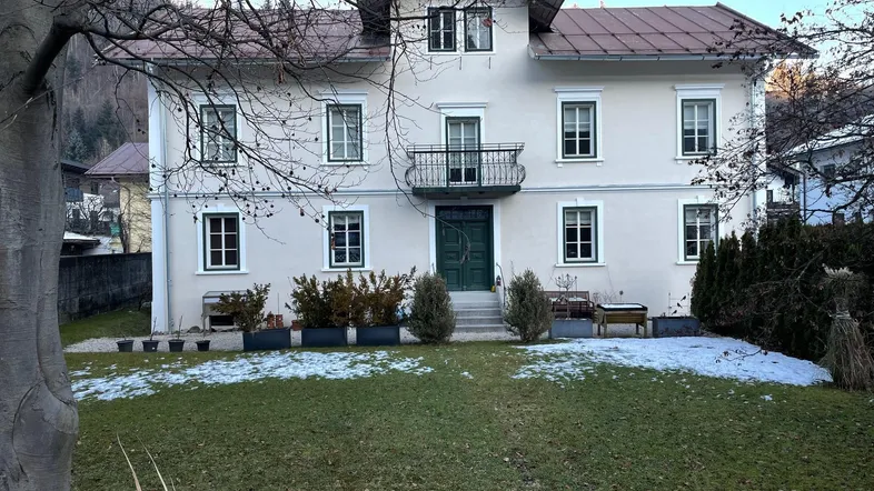 Klassisches Mehrfamilienhaus mit Balkon und Vorgarten in einer bergigen Umgebung.