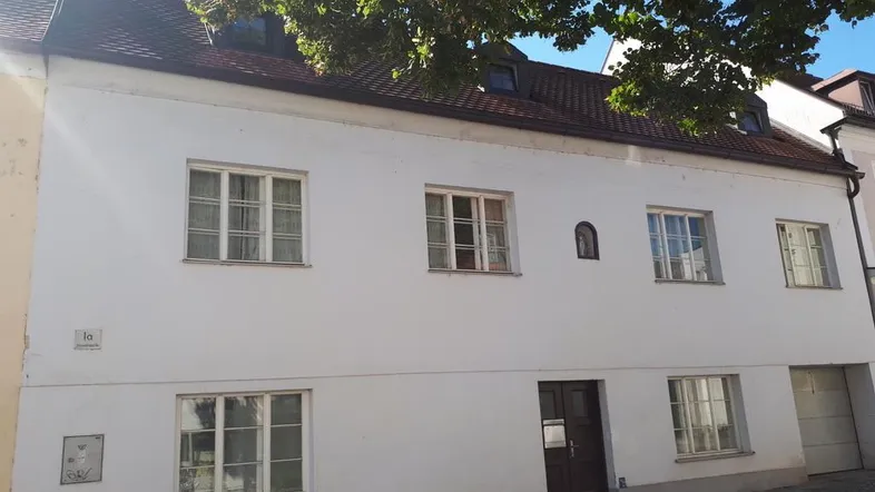 Weißes Mehrfamilienhaus mit mehreren Fenstern und einem roten Ziegeldach in einer städtischen Umgebung.