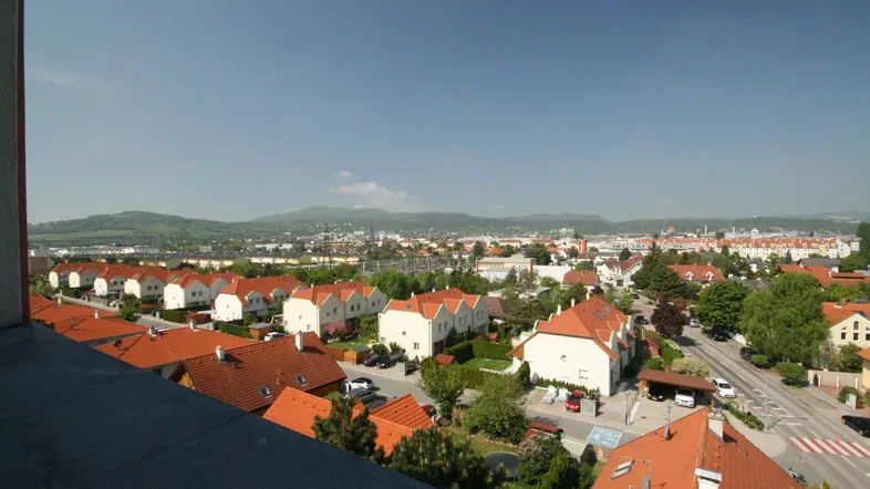 Weitläufiger Blick über eine Wohnsiedlung mit roten Dächern und grünen Hügeln im Hintergrund.