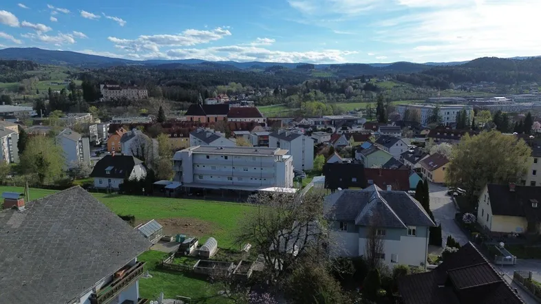 Weitläufiger Blick über die Ortschaft mit Wohngebäuden und grünen Hügeln im Hintergrund unter blauem Himmel.