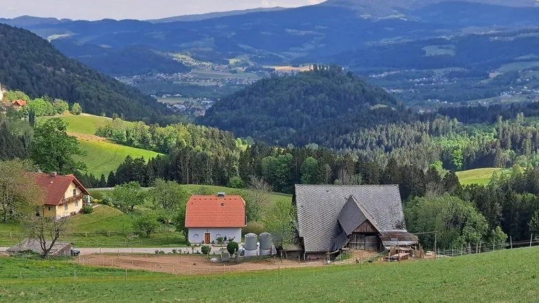 Malerische Landschaftsaufnahme mit Häusern, grünen Hügeln und Wäldern unter bewölktem Himmel.