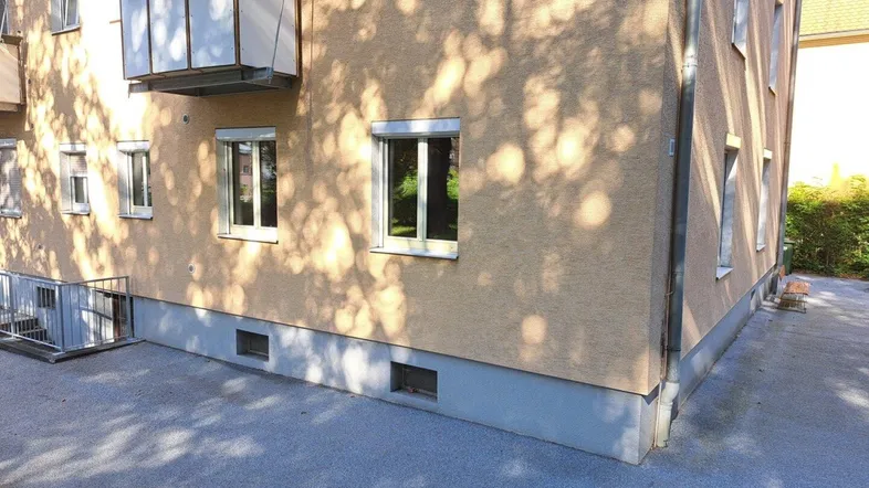 Fassade eines Mehrfamilienhauses mit Balkon und Fenstern, teilweise im Schatten von Bäumen.