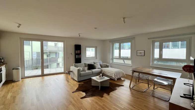 Heller offener Wohn- und Essbereich mit Holzboden, Sofa und Zugang zum Balkon.