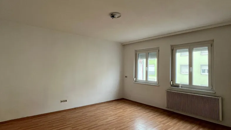 Helles Zimmer mit Laminatboden, weißen Wänden und zwei Fenstern mit Rollläden.
