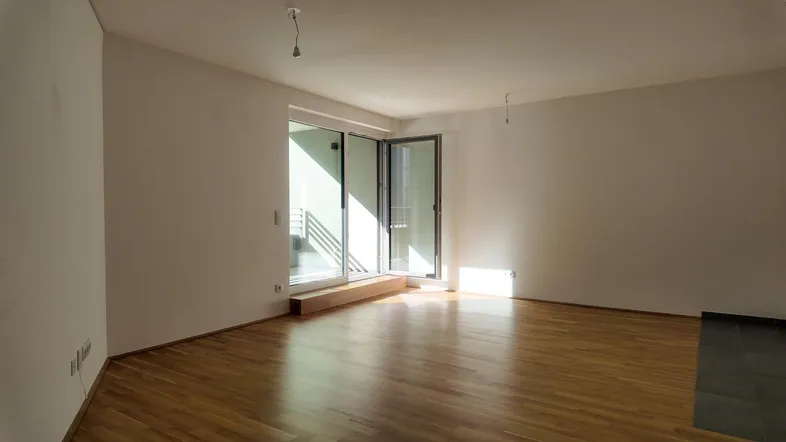 Helles, leeres Zimmer mit Holzboden und großen Schiebetüren, die zu einem Balkon führen.