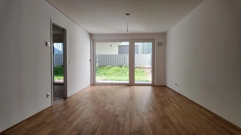 Helles, leeres Zimmer mit Holzboden und großen Fenstern, die den Zugang zum Garten ermöglichen.