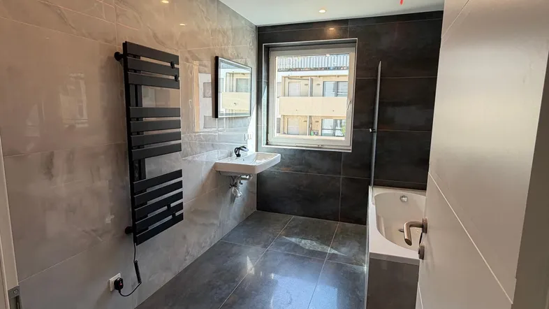Modernes Badezimmer mit grauen Fliesen, Badewanne, Waschbecken und beheiztem Handtuchhalter.