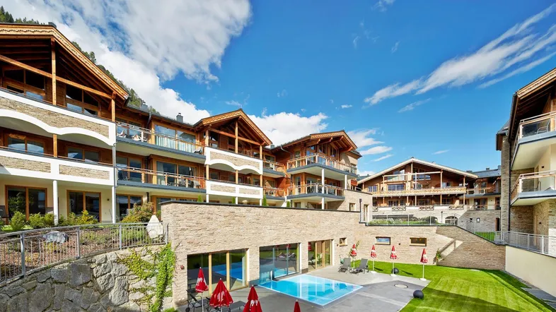 Mehrere Gebäude im alpinen Stil mit Außenpool und gepflegter Grünfläche unter blauem Himmel.