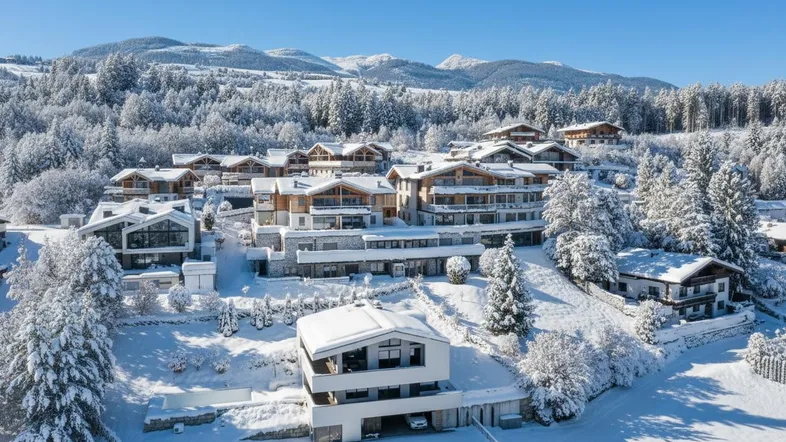Winterlandschaft mit luxuriösen Chalets und verschneiten Bergen im Hintergrund unter blauem Himmel.