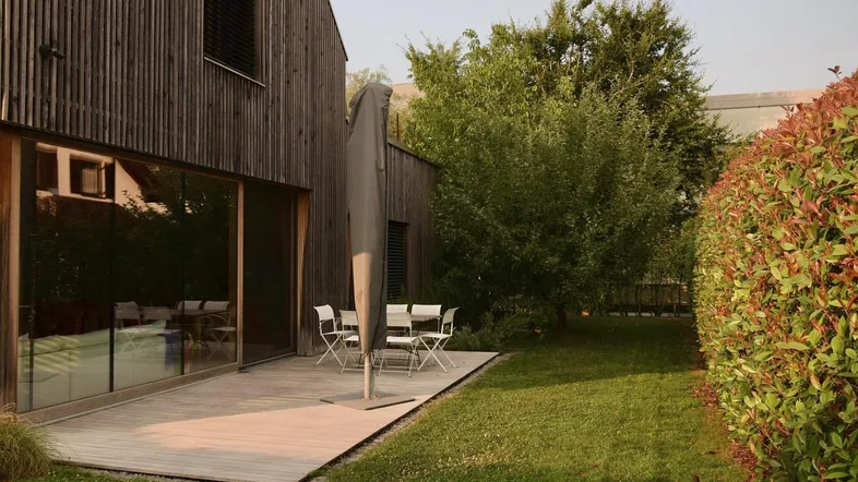 Holzterrasse mit Gartenmöbeln und Sonnenschirm, umgeben von grünem Rasen und Hecken.