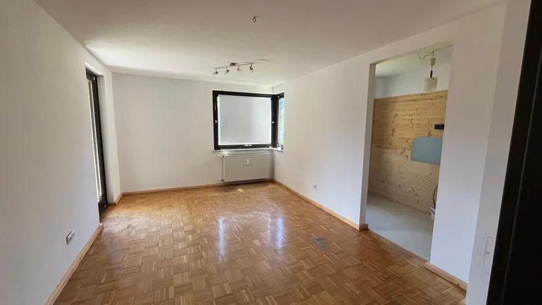 Helles Zimmer mit Parkettboden, weißen Wänden und Fenster, das zu einem holzvertäfelten Raum führt.