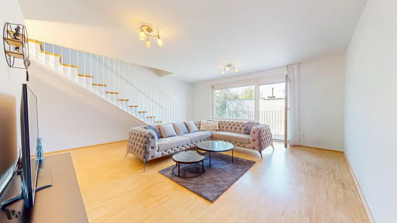 Helles Wohnzimmer mit beigem Ecksofa, Holzboden und Zugang zum Balkon über eine Glastür.