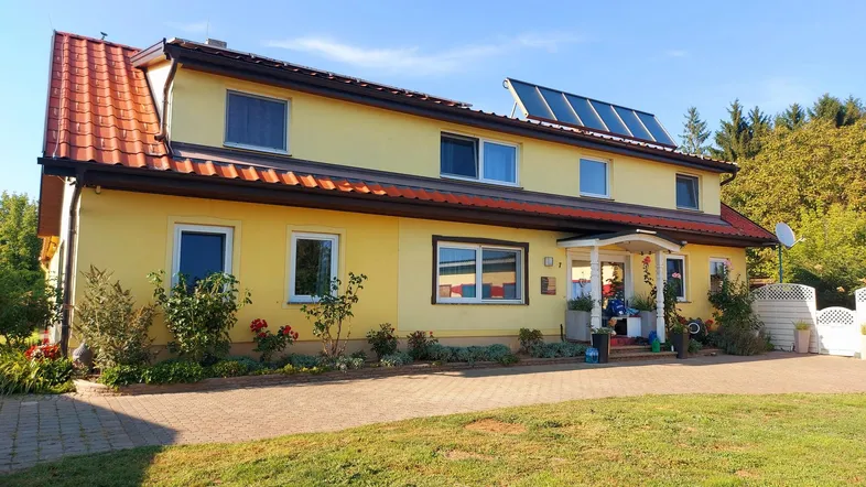 Gelbes Wohnhaus mit rotem Ziegeldach, Solaranlage und gepflegtem Vorgarten unter blauem Himmel.