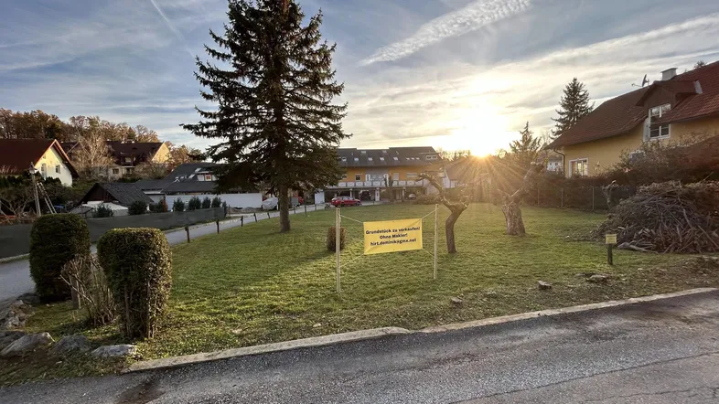 Sonniges Grundstück mit Verkaufsanzeige, umgeben von Bäumen und Häusern, unter blauem Himmel.