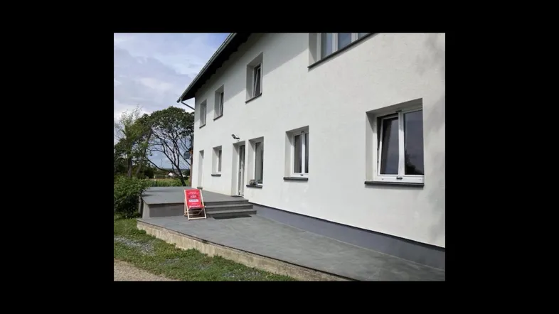 Seitenansicht des weißen Gebäudes mit grauen Akzenten und umgebender Natur, unter blauem Himmel.