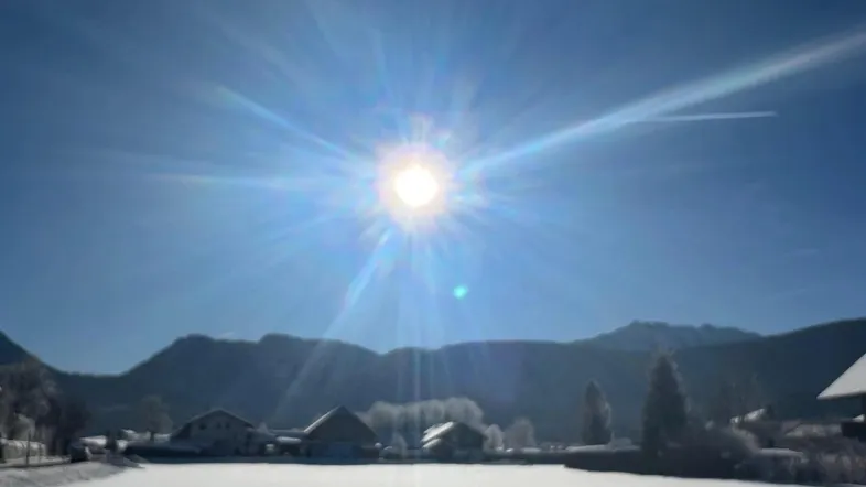 Winterlandschaft mit schneebedeckten Feldern, Häusern und strahlendem Sonnenschein vor einer Bergkulisse.