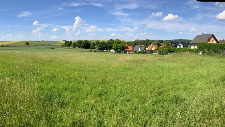 Weitläufiges grünes Feld mit ländlicher Bebauung im Hintergrund unter blauem Himmel.