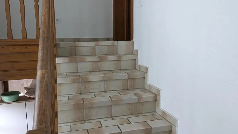 Geflieste Treppe mit Holzgeländer und weißen Wänden, die in den oberen Bereich führt.