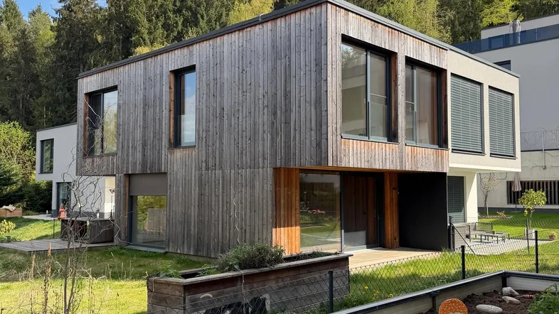 Modernes Wohnhaus mit Holzfassade und großen Fenstern, umgeben von grüner Natur und Bäumen.