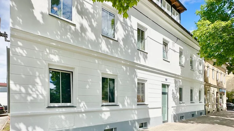Weißes Mehrfamilienhaus mit mehreren Fenstern und einem Dachgeschossausbau unter blauem Himmel.