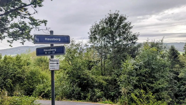 Wegweiser zu Hausberg mit Hinweisschild für Privatweg, umgeben von üppiger Vegetation und bewölktem Himmel.