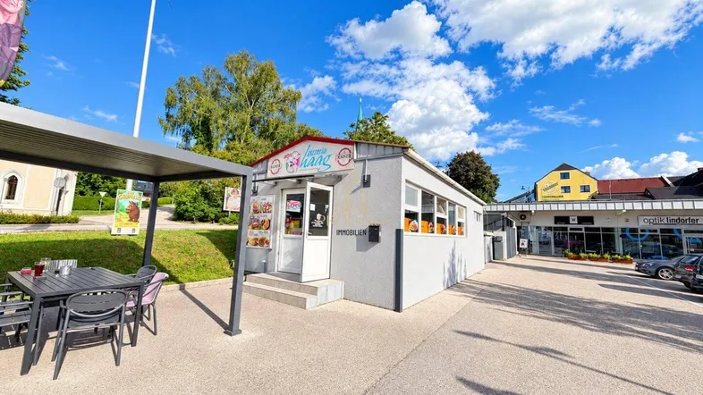 Kleines Gewerbegebäude mit überdachtem Außenbereich und Sitzgelegenheiten, umgeben von einem Parkplatz.