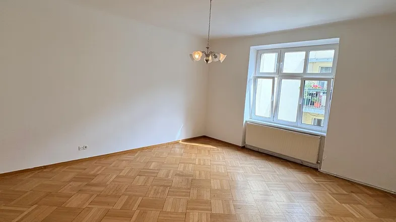 Helles, leeres Wohnzimmer mit klassischem Parkettboden und einem großen Fenster für viel Tageslicht.