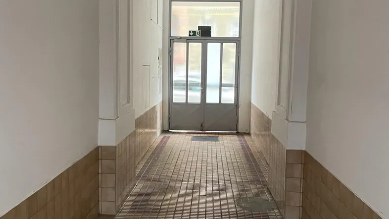 Langer, schmaler Flur mit hellen Wänden, gefliestem Boden und hoher Decke, der zum Ausgang führt.