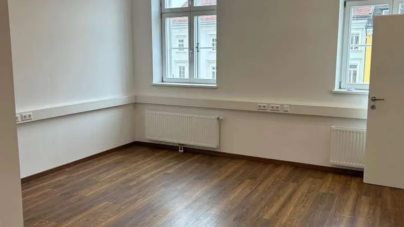 Heller, leerer Raum mit zwei Fenstern, Holzboden und Heizkörpern, ideal als Büro oder Arbeitszimmer.