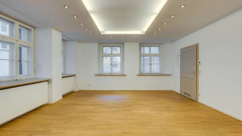 Geräumiges Wohnzimmer mit hellem Holzfußboden, großen Fenstern und moderner Deckenbeleuchtung, in gutem Zustand.
