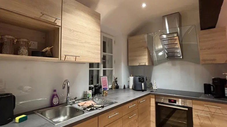 Moderne Küche mit hellen Holzfronten, dunkler Arbeitsplatte, integrierten Geräten und sichtbaren Deckenbalken.