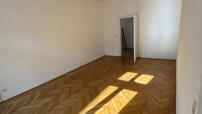 Helles, frisch renoviertes Zimmer mit Fischgrätparkett und weißen Wänden, durchflutet von natürlichem Sonnenlicht.