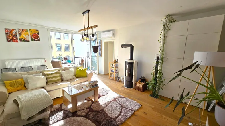 Helles Wohnzimmer mit Holzboden, Kaminofen und Zugang zu einem kleinen Balkon mit Stadtblick.