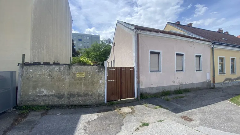 Ansicht eines älteren Hauses mit Putzfassade, einer Mauer und einem Holztor zur Straße hin.