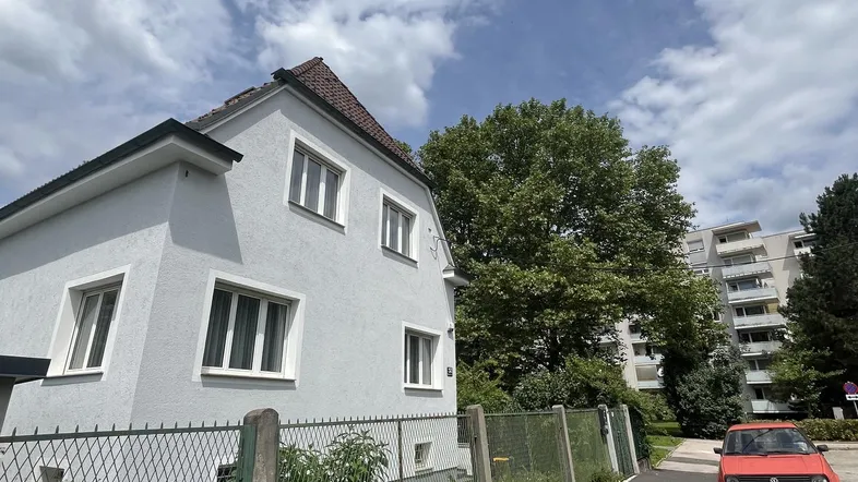 Helles Einfamilienhaus mit gepflegtem Vorgarten und Blick auf die Umgebung unter blauem Himmel.