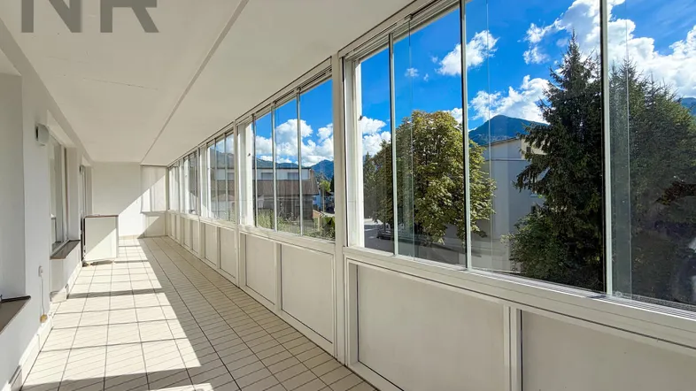 Langer, verglaster Balkon mit Fliesenboden und Blick auf Bäume und Berge.