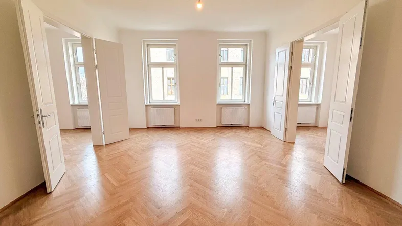 Helles, leeres Wohnzimmer mit Fischgrätparkettboden und hohen Doppeltüren zu angrenzenden Räumen.