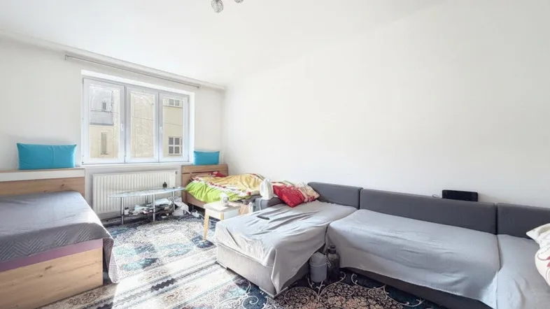 Heller Wohnbereich mit großem Sofa, zwei Betten und einem farbenfrohen Teppichboden.