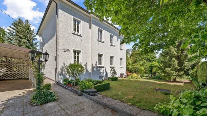 Weißes Wohnhaus mit gepflegtem Garten, großer Rasenfläche, altem Baumbestand und gepflasterter Zufahrt.