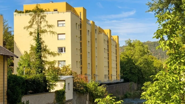 Gelbes Mehrfamilienhaus am Flussufer, umgeben von üppigem Grün unter blauem Himmel.