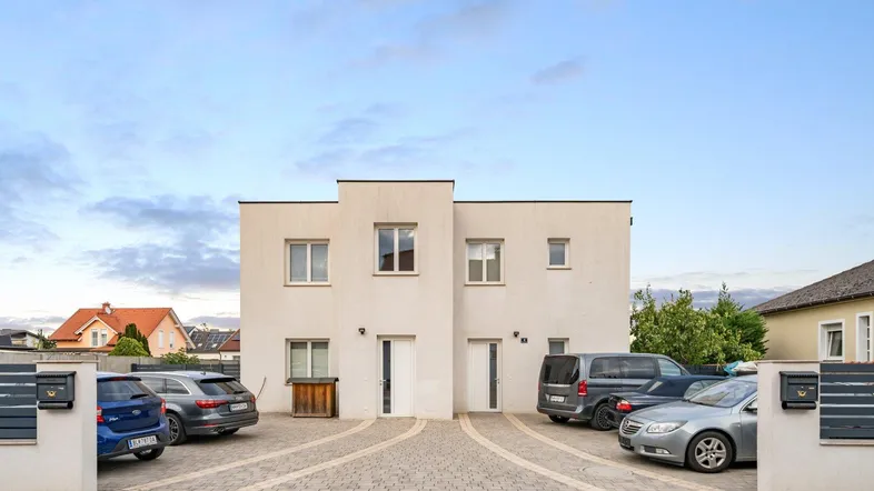 Moderne weiße Doppelhaushälfte mit gepflastertem Vorplatz und Parkplätzen für mehrere Fahrzeuge.
