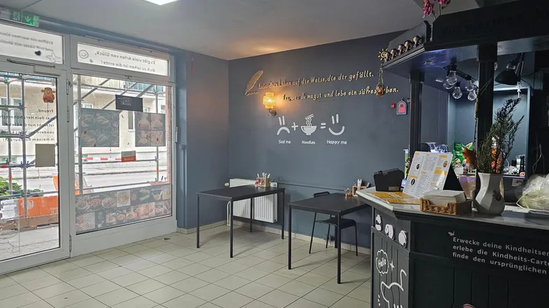Innenansicht eines kleinen Restaurants oder Imbisses mit Theke und Sitzgelegenheiten. Große Fensterfront zur Straße.