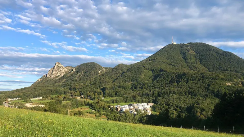 Weitläufige Landschaft mit grünen Wiesen, bewaldeten Bergen und einem kleinen Dorf unter blauem Himmel.
