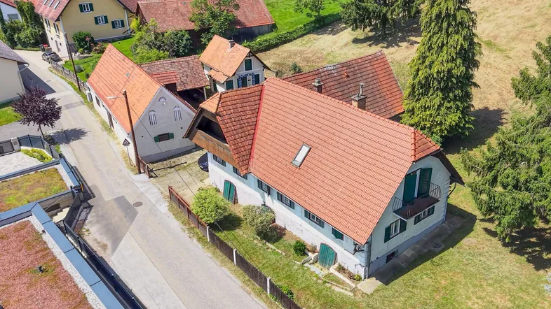 Drohnenaufnahme eines traditionellen Hauses mit rotem Ziegeldach und umliegenden Gebäuden in ländlicher Umgebung.