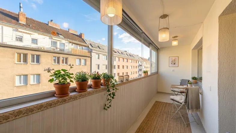 Heller, verglaster Balkon mit Sitzgelegenheiten und Pflanzen, bietet einen Blick auf die umliegenden Gebäude.