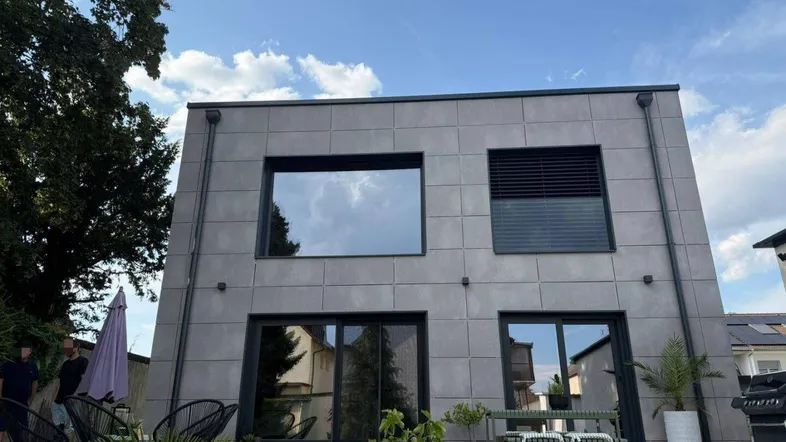 Moderne Hausfassade mit großen Fenstern, Flachdach und kleiner Terrasse im Vordergrund.