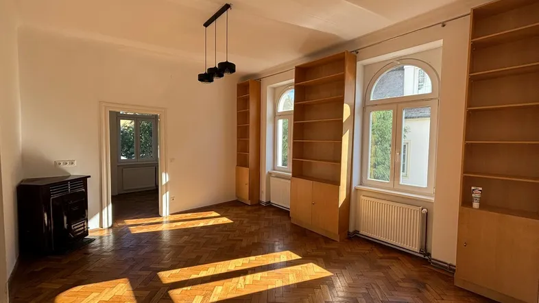 Helles Wohnzimmer mit Fischgrätparkett, Einbauregalen und einem Ofen, durchflutet von Sonnenlicht.