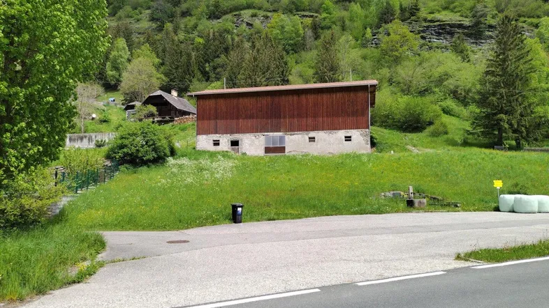 Ländliches Gebäude mit Holzwänden und Steinfundament, umgeben von grünen Hügeln und Wald.